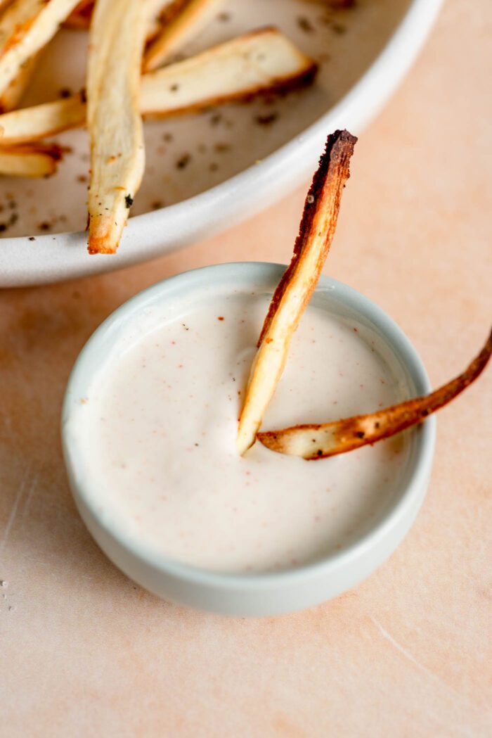 Two parsnip fries being dipped in a small dish of mayo.