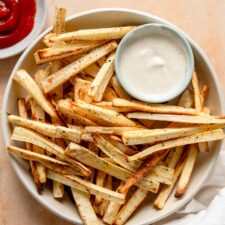 Plate of baked parsnip fries with a small dish of dip and a dish of ketchup beside the plate.