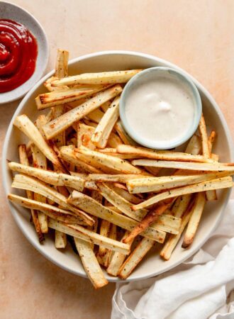 Plate of baked parsnip fries with a small dish of dip and a dish of ketchup beside the plate.