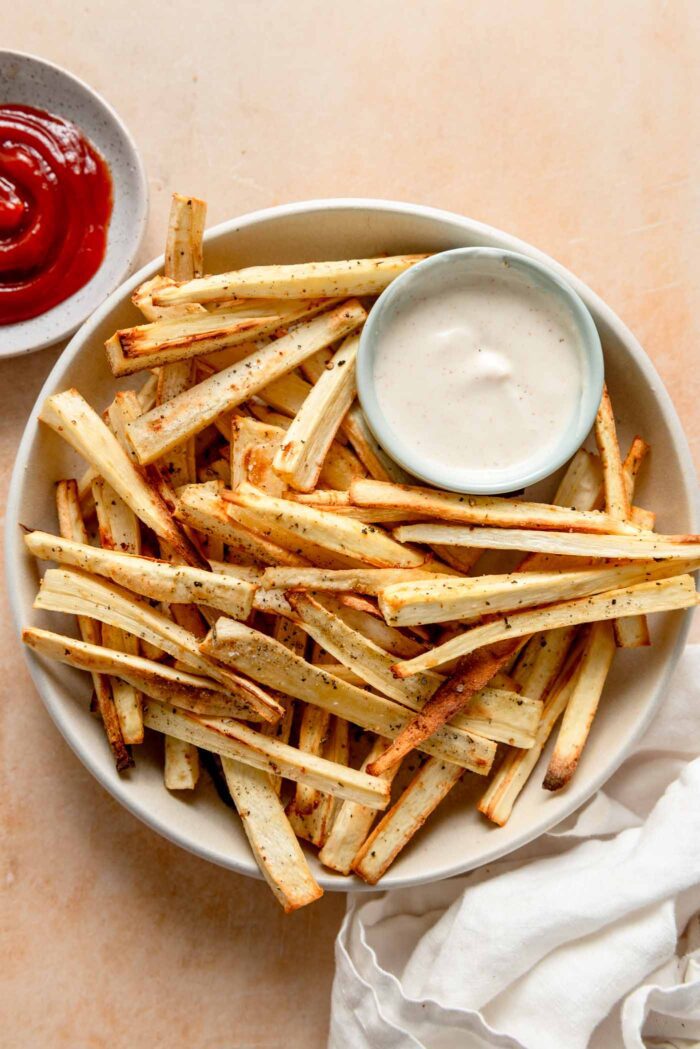 Plate of baked parsnip fries with a small dish of dip and a dish of ketchup beside the plate.