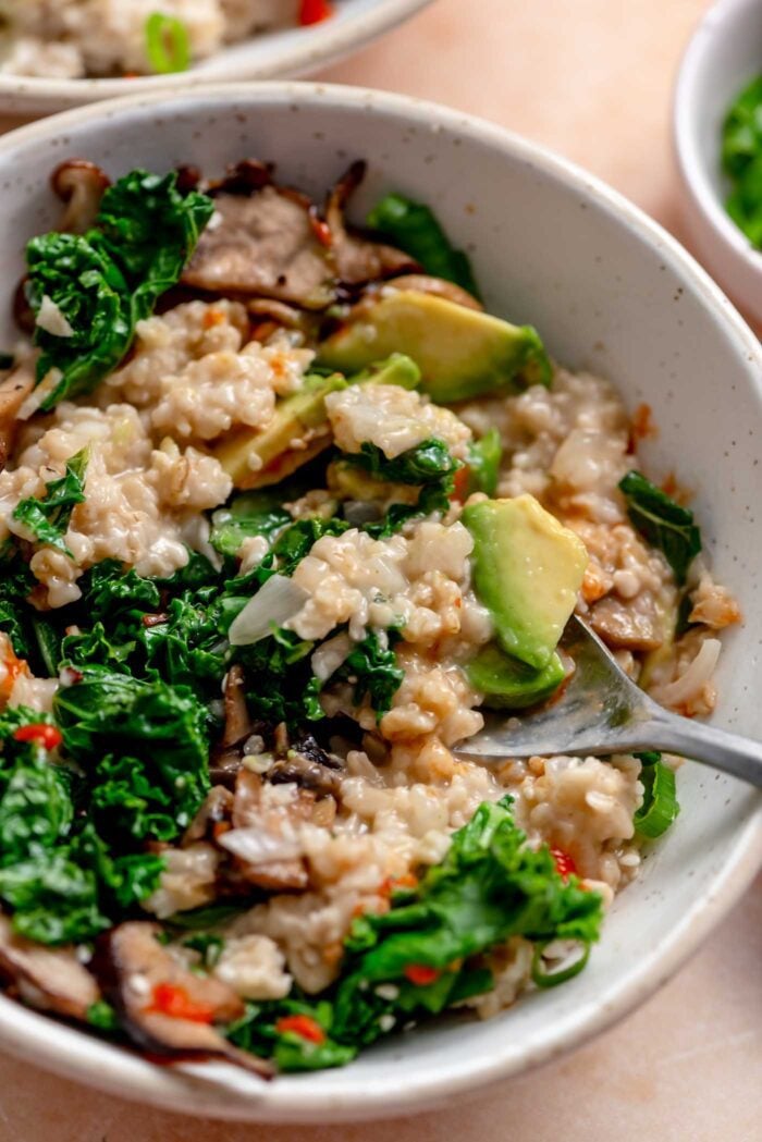 Spoon in a bowl of savory oatmeal mixed with kale, mushroom, avocado and hot sauce.