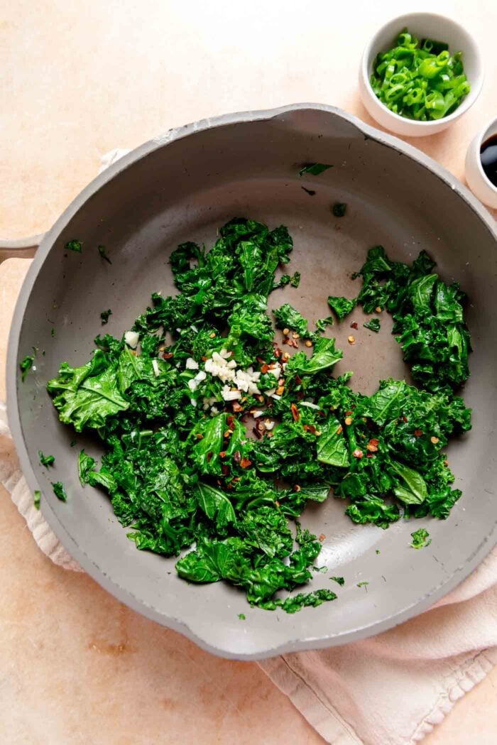 Cooked chopped kale sprinkled with chili flakes and minced garlic in a skillet.