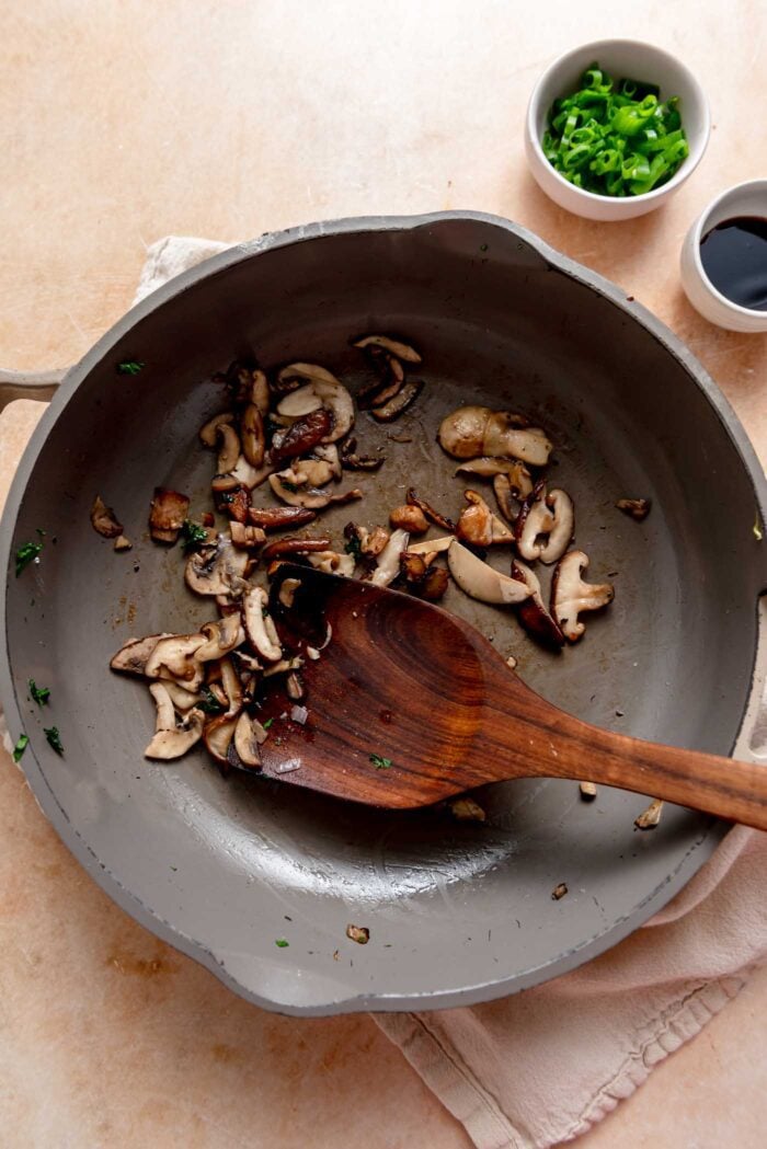 Thinly sliced sauteed mushrooms in a skillet with a wooden spoon.