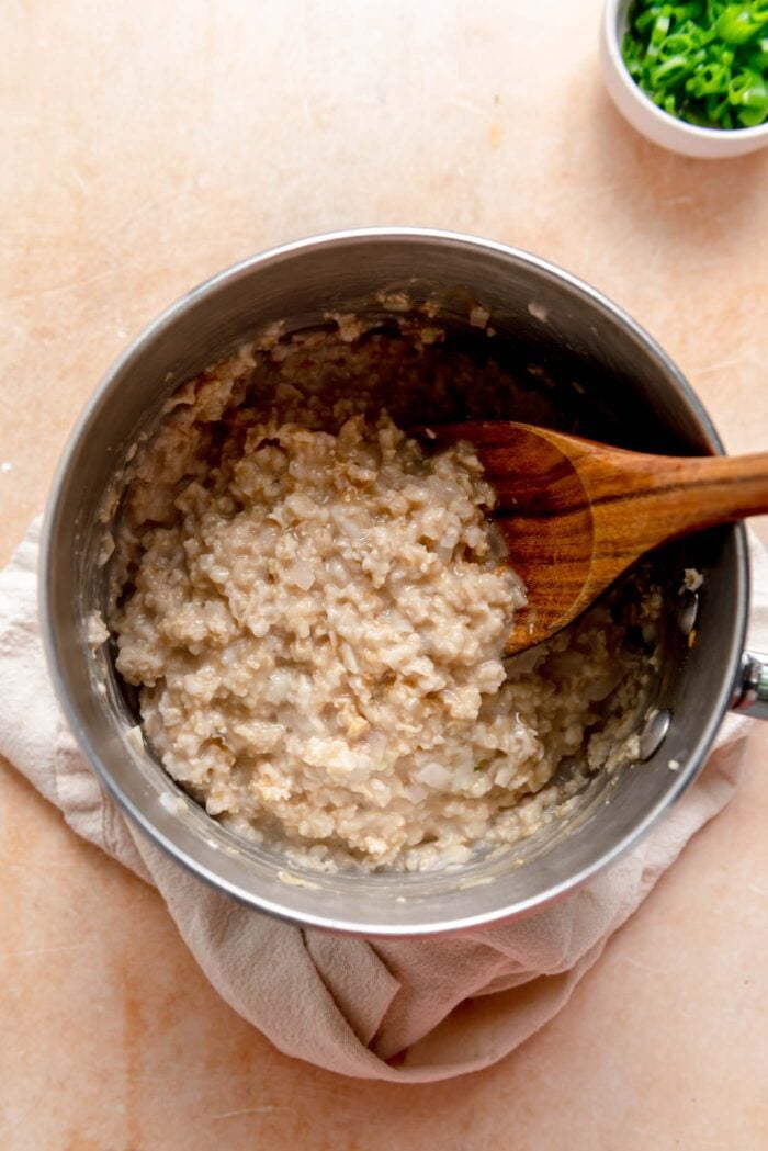 Creamed cooked oats in a metal saucepan with a wooden spoon resting in it.