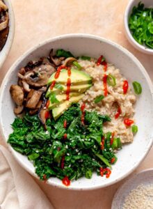 Bowl of savory oatmeal topped with cooked kale, thinly sliced mushrooms, sliced avocado and a drizzle of hot sauce.