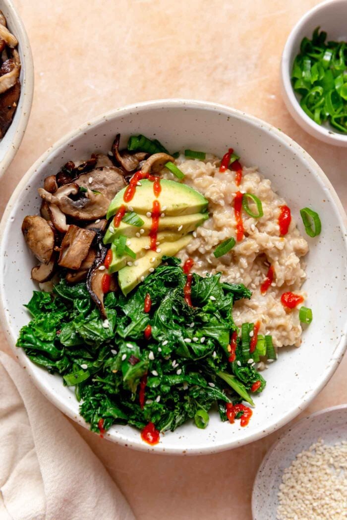 Bowl of savory oatmeal topped with cooked kale, thinly sliced mushrooms, sliced avocado and a drizzle of hot sauce.