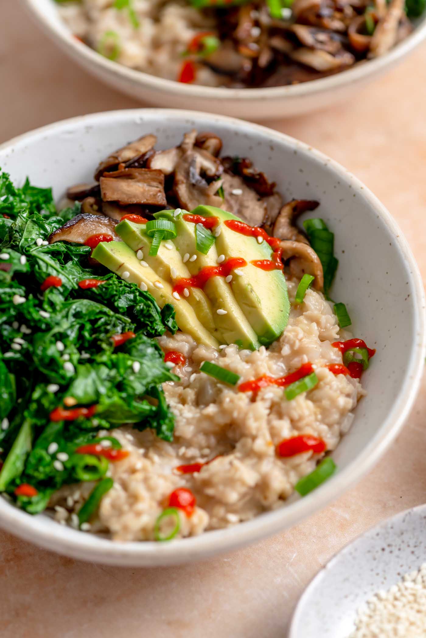 Bowl of savory oatmeal topped with avocado slices, cooked kale and mushroom and some hot sauce.