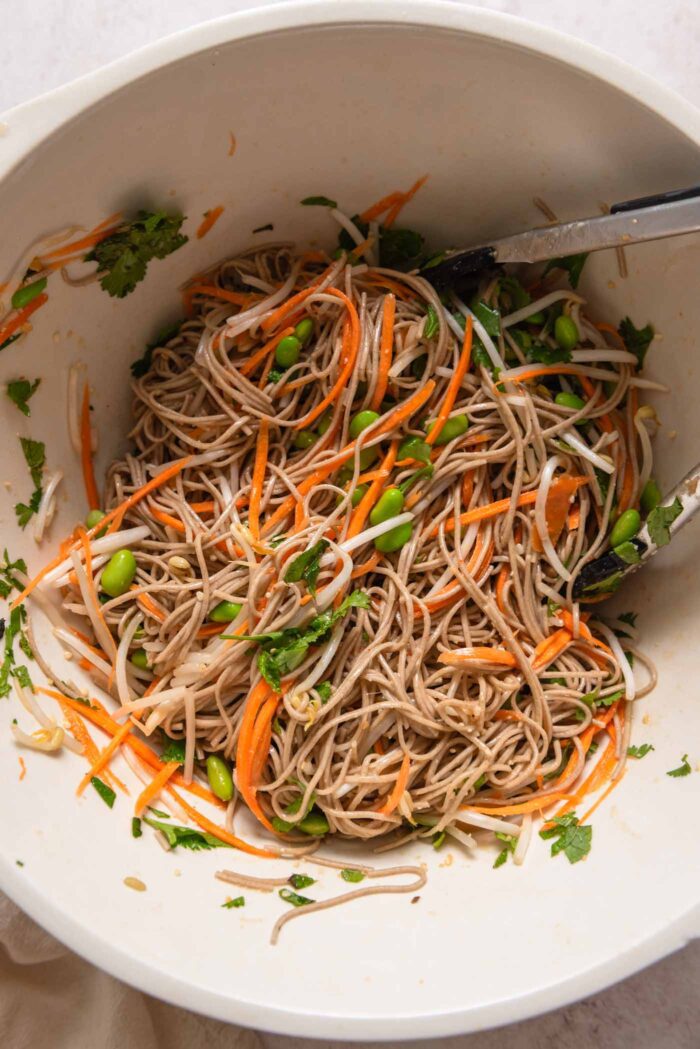 Cold soba noodle salad with edamame, carrot and bean sprouts in a large mixing bowl with a pair of tongs resting in it.