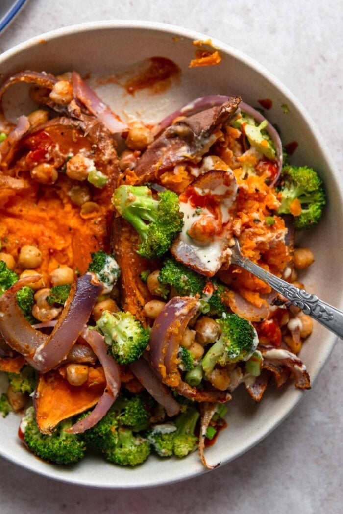 Mashed sweet potato with hot sauce, tahini sauce, broccoli, chickpeas and sliced red onion on a plate.