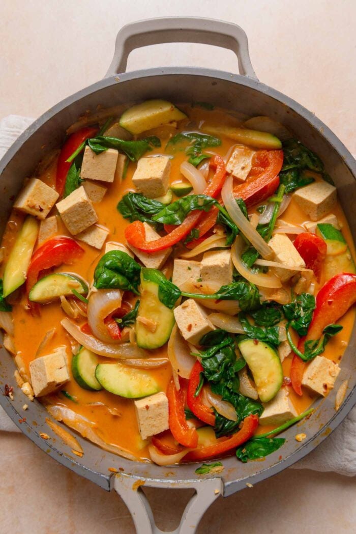 Red curry with bell pepper, spinach, tofu, onion and zucchini cooking in a pan.