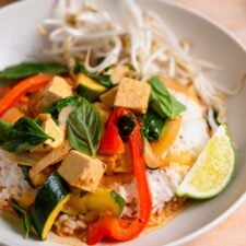 Bowl of Thai red curry with cubes of tofu, bell pepper and zucchini with fresh bean sprouts, basil leaves and a lime wedge.