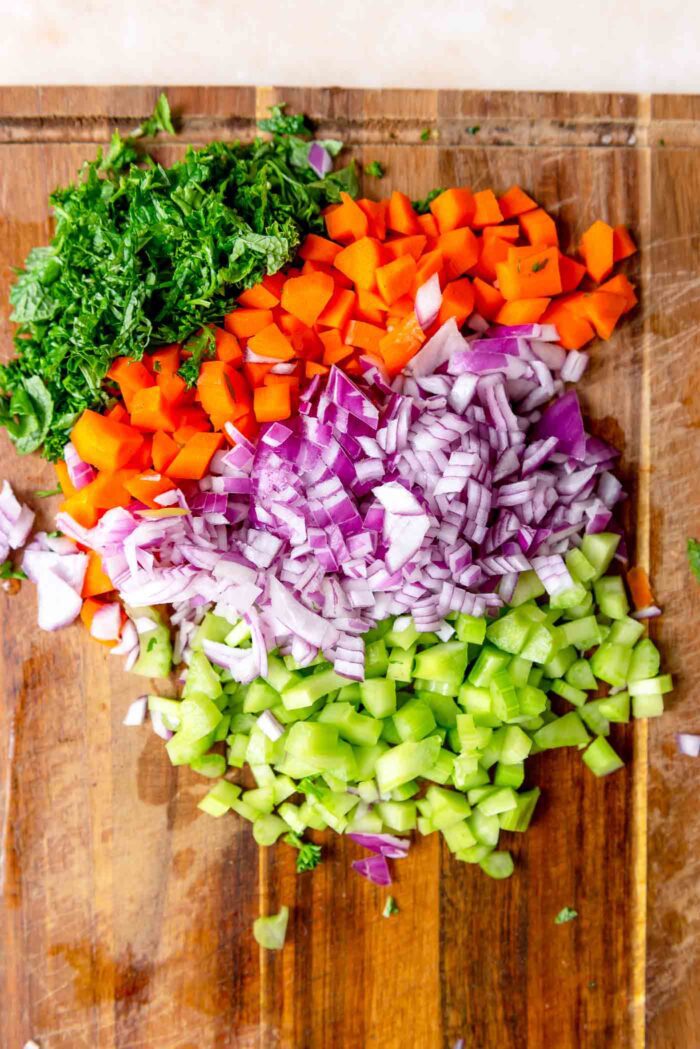 Chopped herbs, onion, carrot and celery on a wooden cutting board.