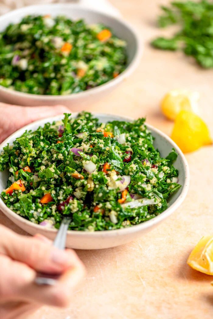 Hands using a fork in a bowl of kale quinoa salad with some wedges of lemon in the background.