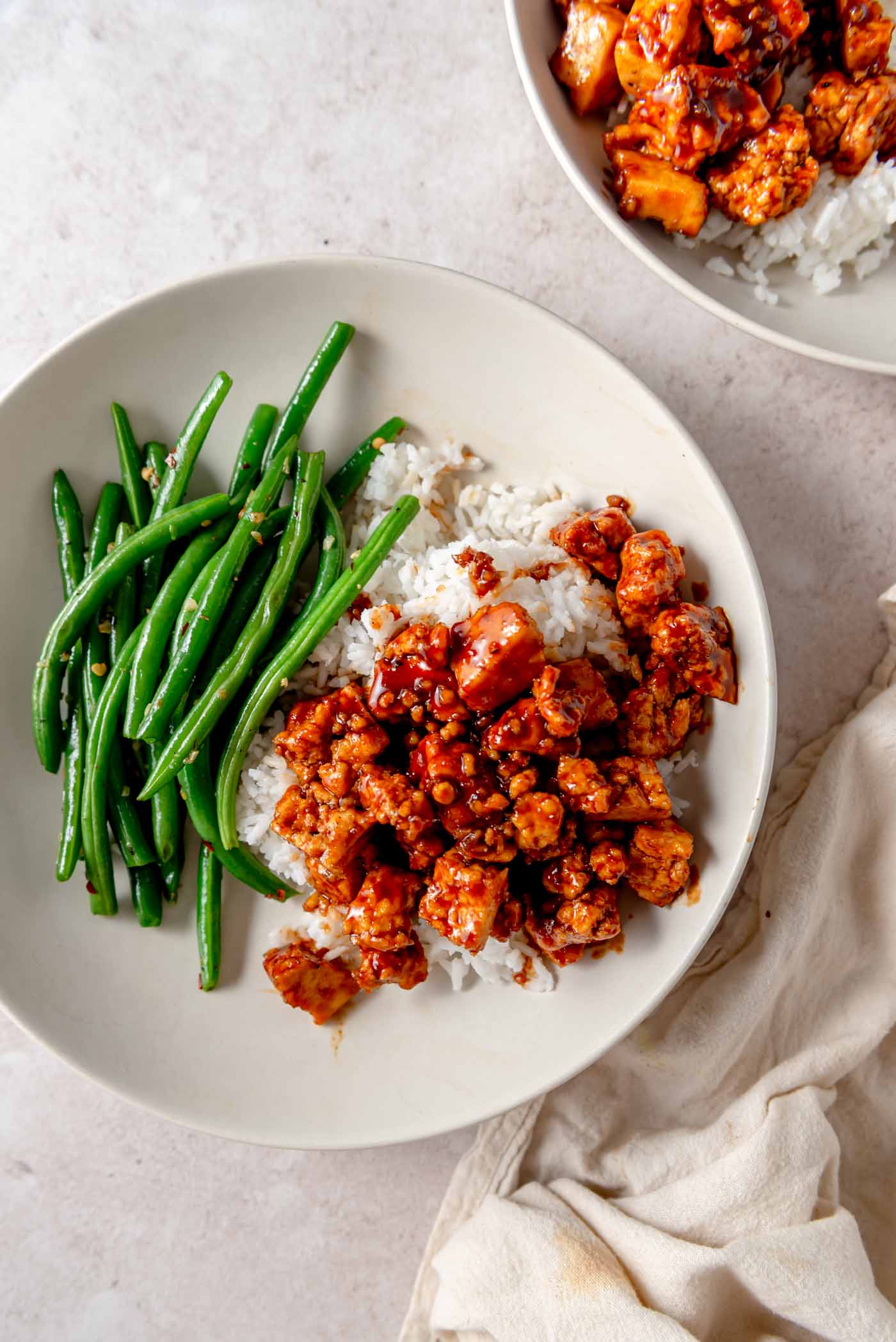 Tofu cubes tossed in a gochujang sauce over rice with a serving of green beans in a bowl.