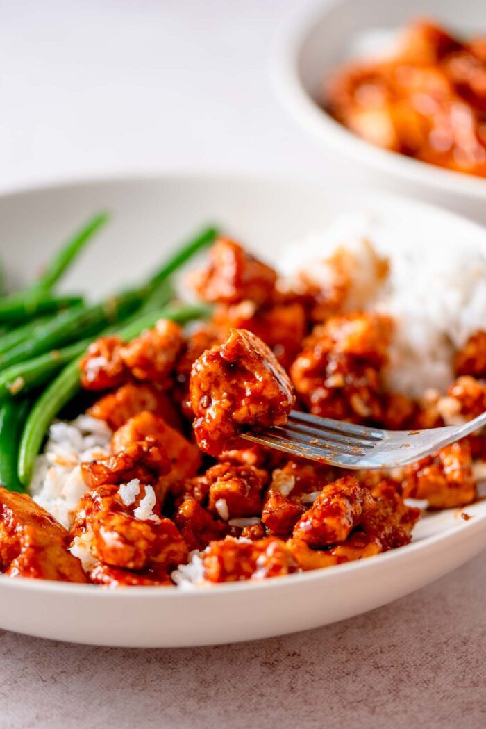 A piece of gochujang tofu on a fork over a bowl of more tofu, rice and green beans.