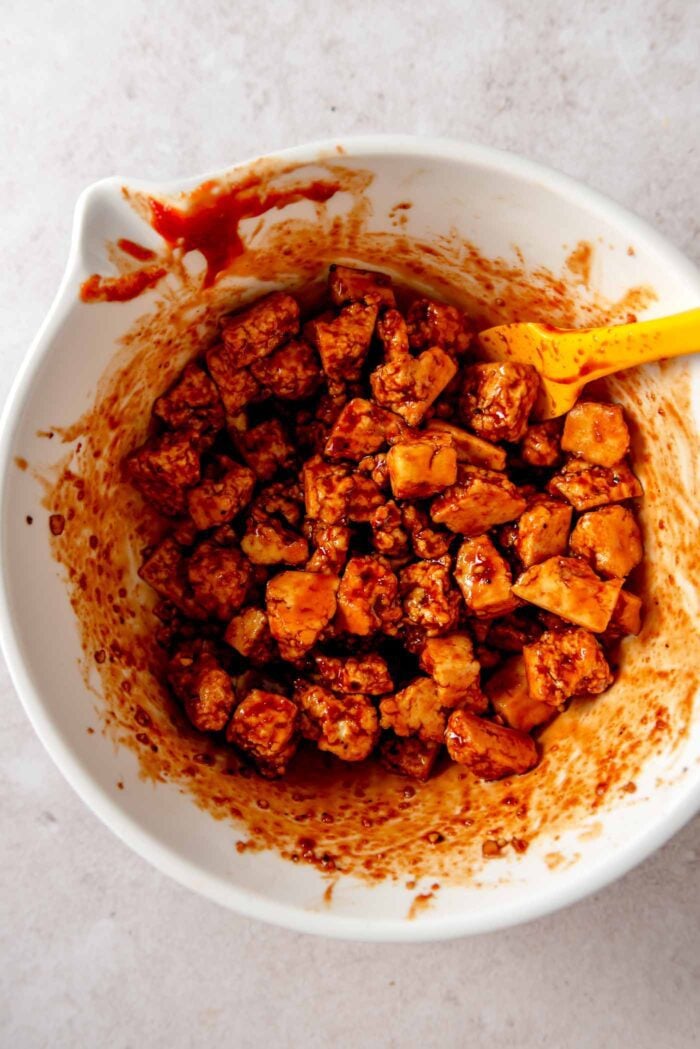 Bite-sized pieces of tofu tossed in a glaze sauce a mixing bowl with a small rubber spatula in it.