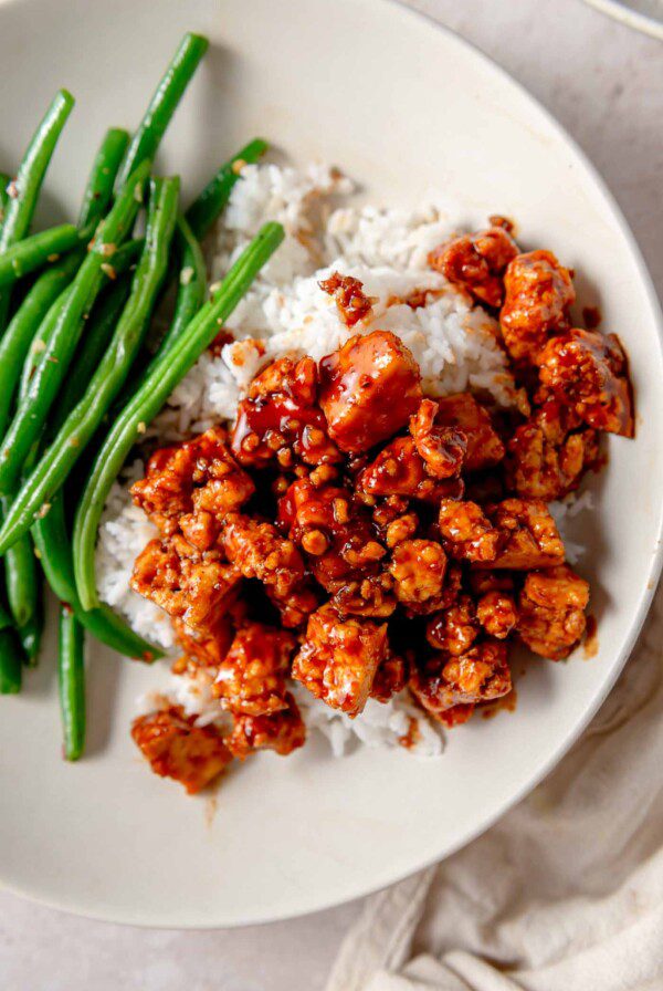 Tofu cubes tossed in a gochujang glaze over rice with a serving of green beans in a bowl.