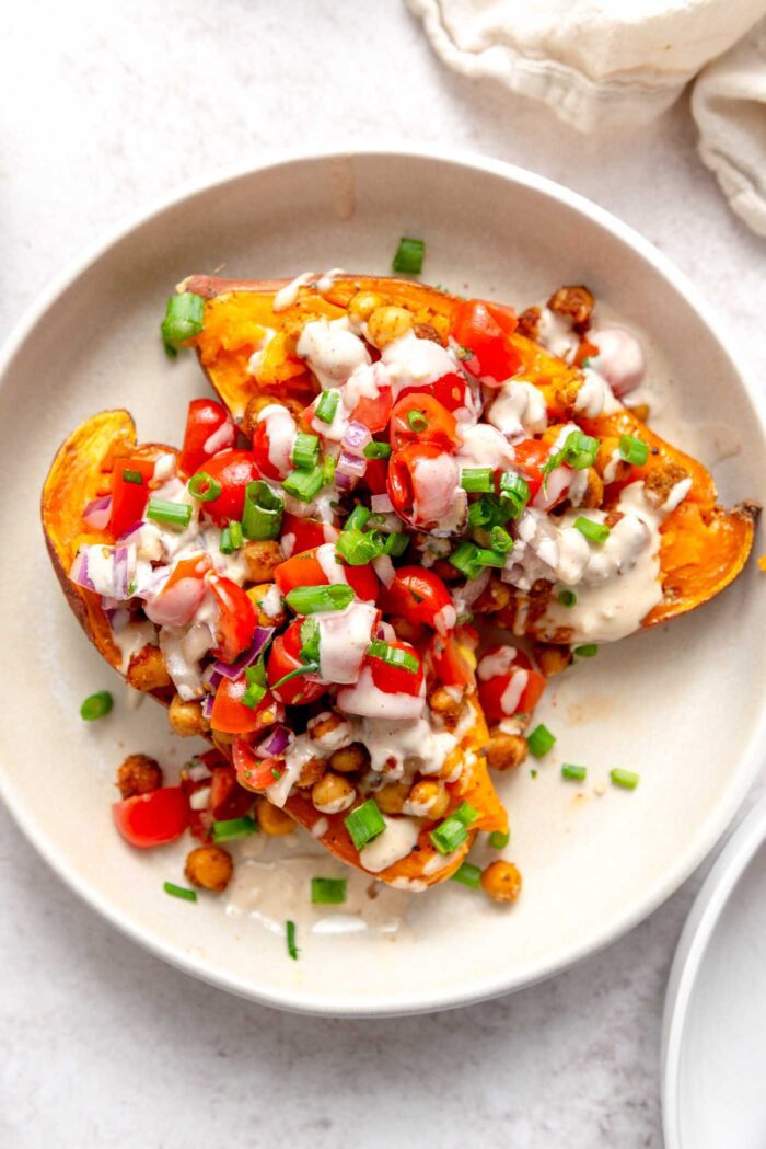 Two halves of a baked sweet potato topped with roasted chickpeas, tomatoes, parsley and creamy tahini sauce on plate.