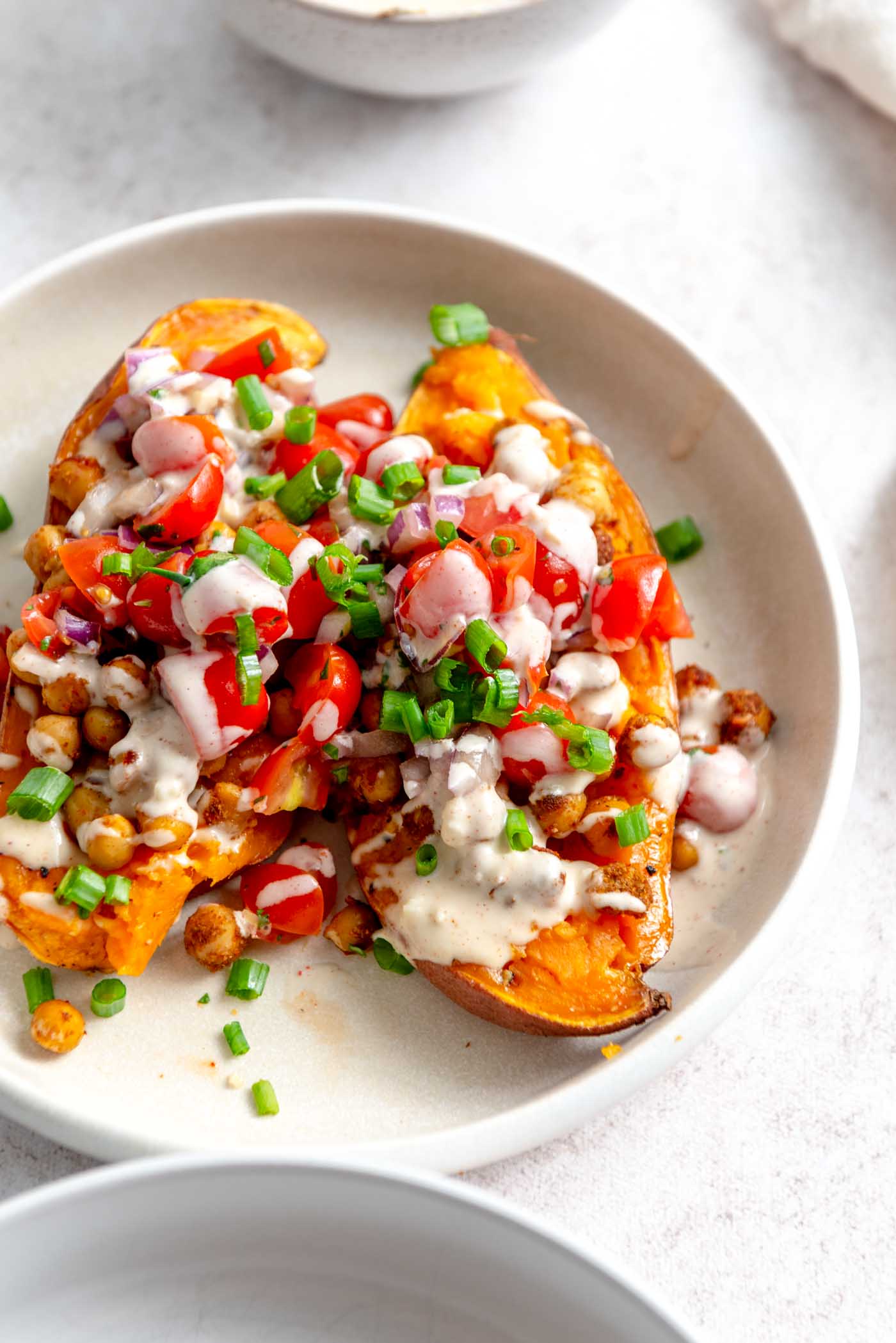 Two baked sweet potato halves topped with chickpeas, cherry tomatoes, parsley and tahini sauce.