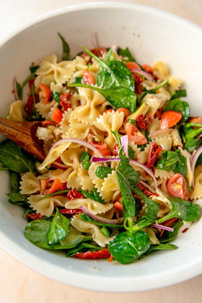 Sun dried tomato pasta salad with fresh spinach, red onion, cherry tomatoes and artichokes in a mixing bowl.