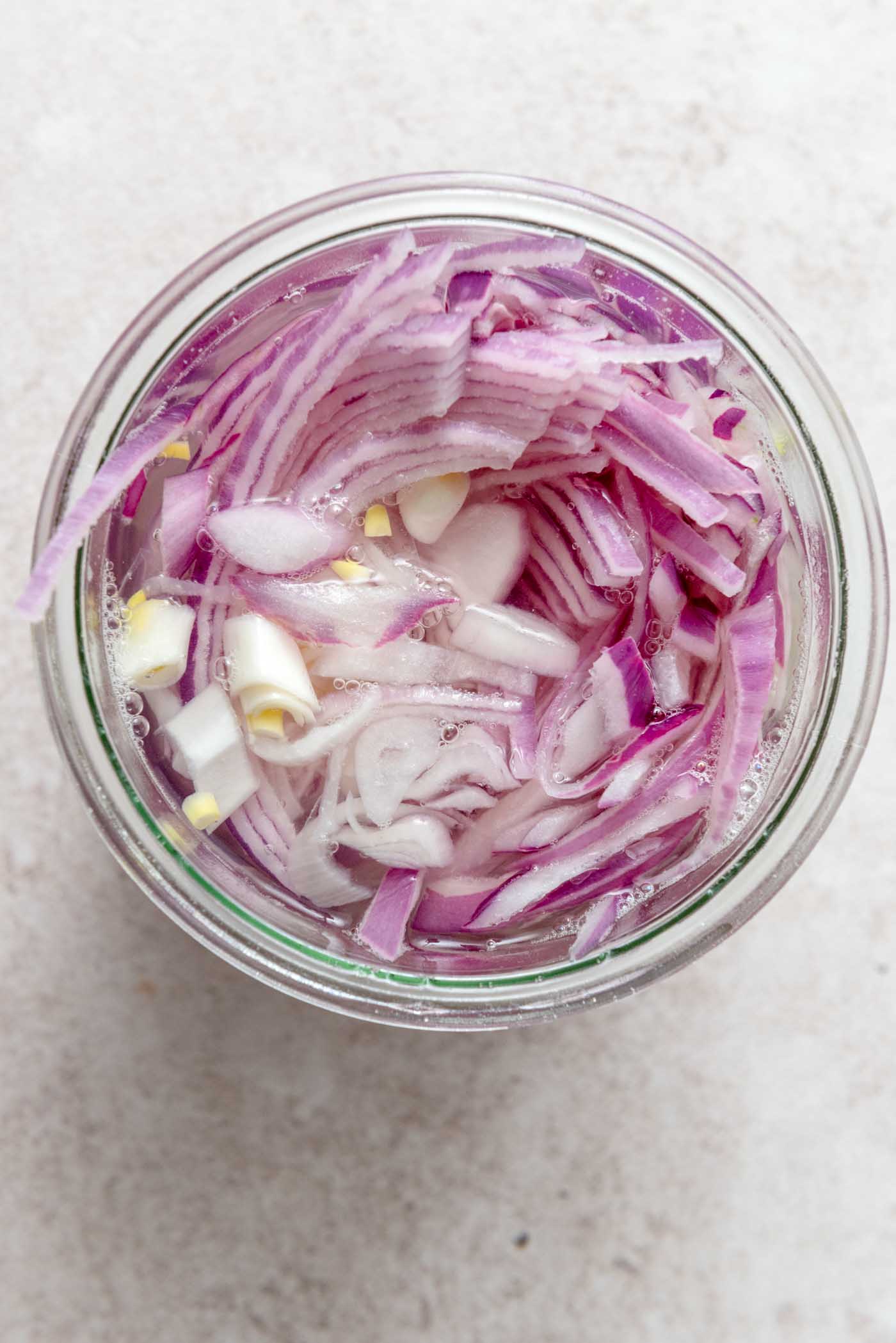 Thinly sliced red onions submerged in a clear liquid within a glass jar, beginning the pickling process.