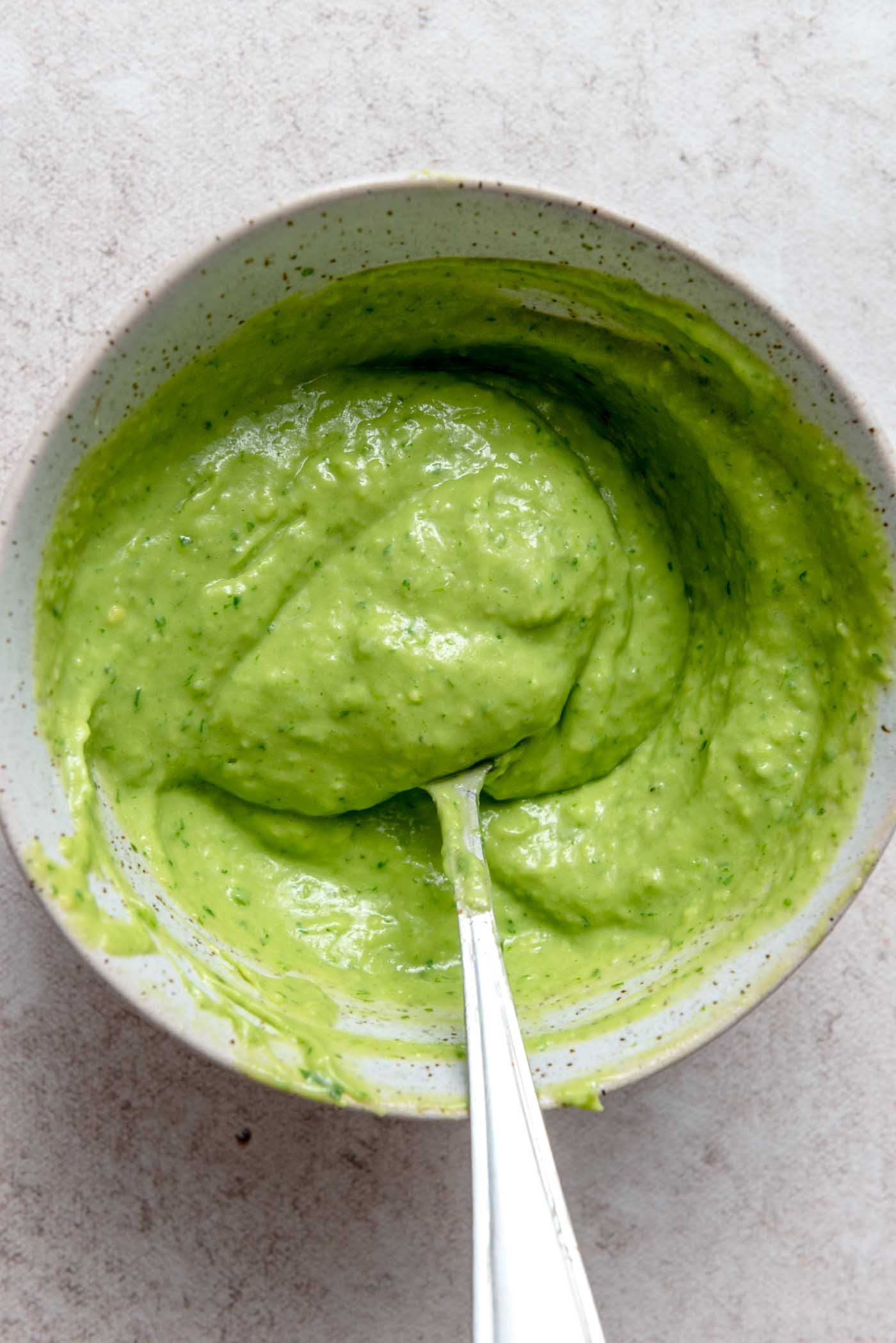 A smooth, vibrant green creamy avocado cilantro lime sauce in a small bowl, with a spoon resting in it, ready to be served.