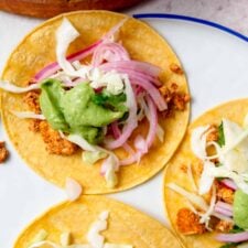 Three tofu tacos topped with cabbage, avocado sauce and pickled onion on a plate.