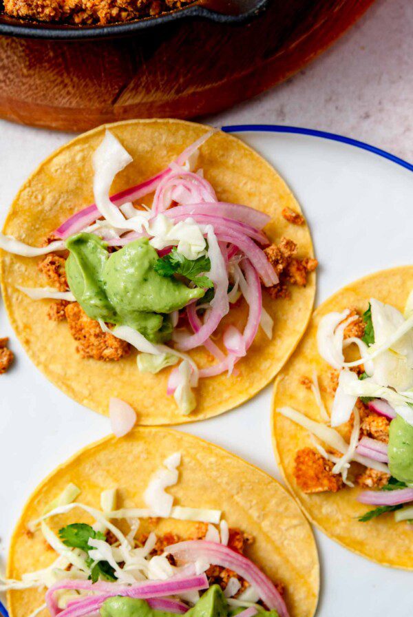 Three tofu tacos topped with cabbage, avocado sauce and pickled onion on a plate.