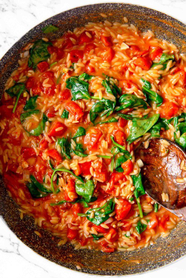 Tomato orzo with spinach cooking in a pan with a spoon resting in it.