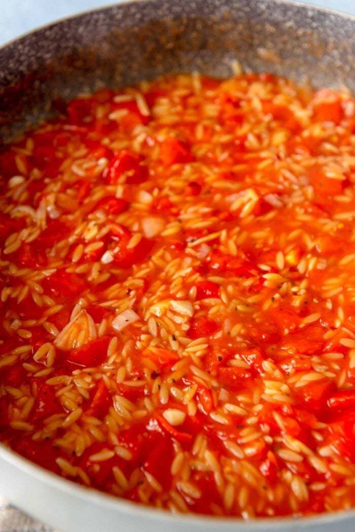 Orzo cooking in a tomato sauce in a large pan.