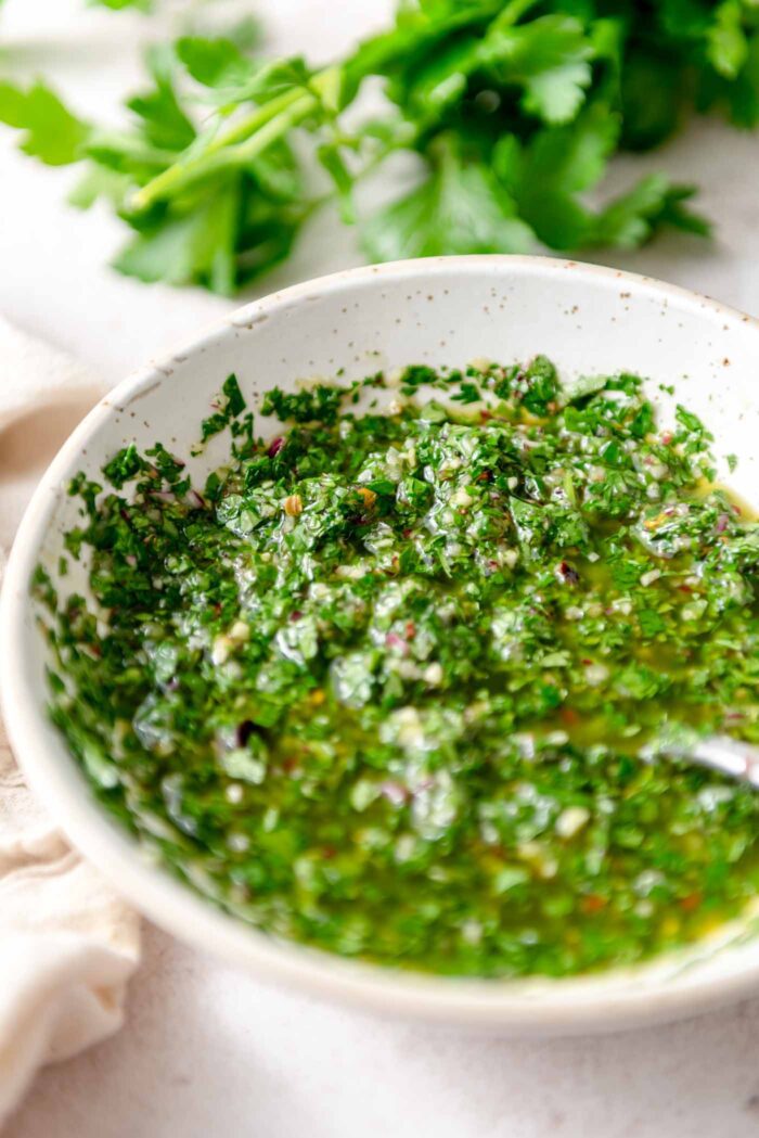 Bowl of chimmichurri sauce with a spoon in it and some fresh herbs in the background.