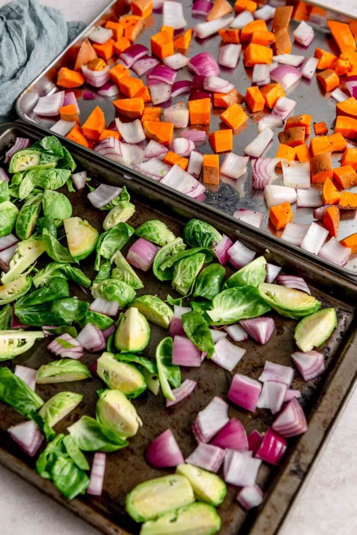 Two baking sheets with chopped brussels sprouts, sweet potato and red onion.