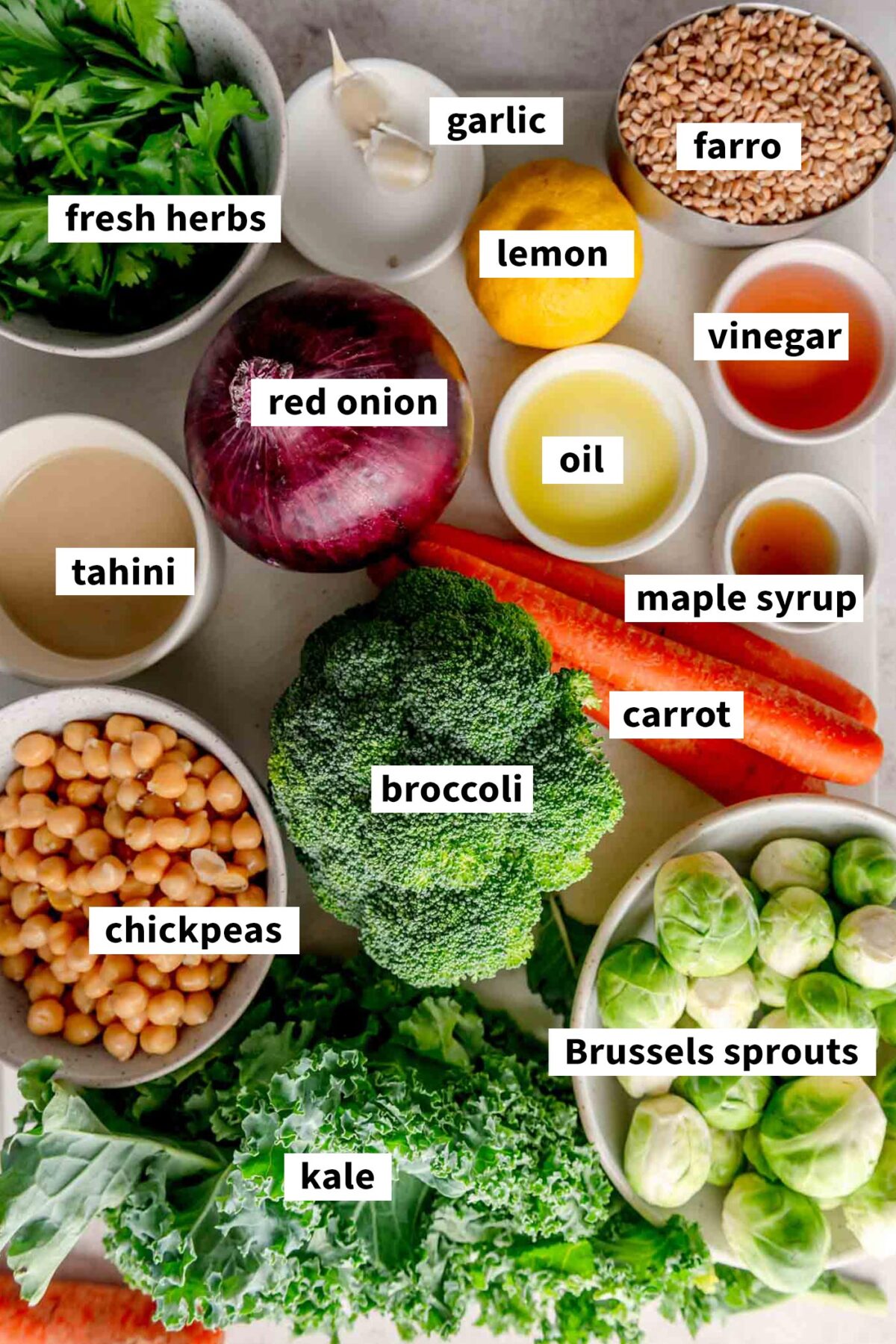 All of the ingredients for making a vegan farro bowl with tahini sauce, roasted vegetables and roasted chickpeas on a cutting board and each labelled with text.