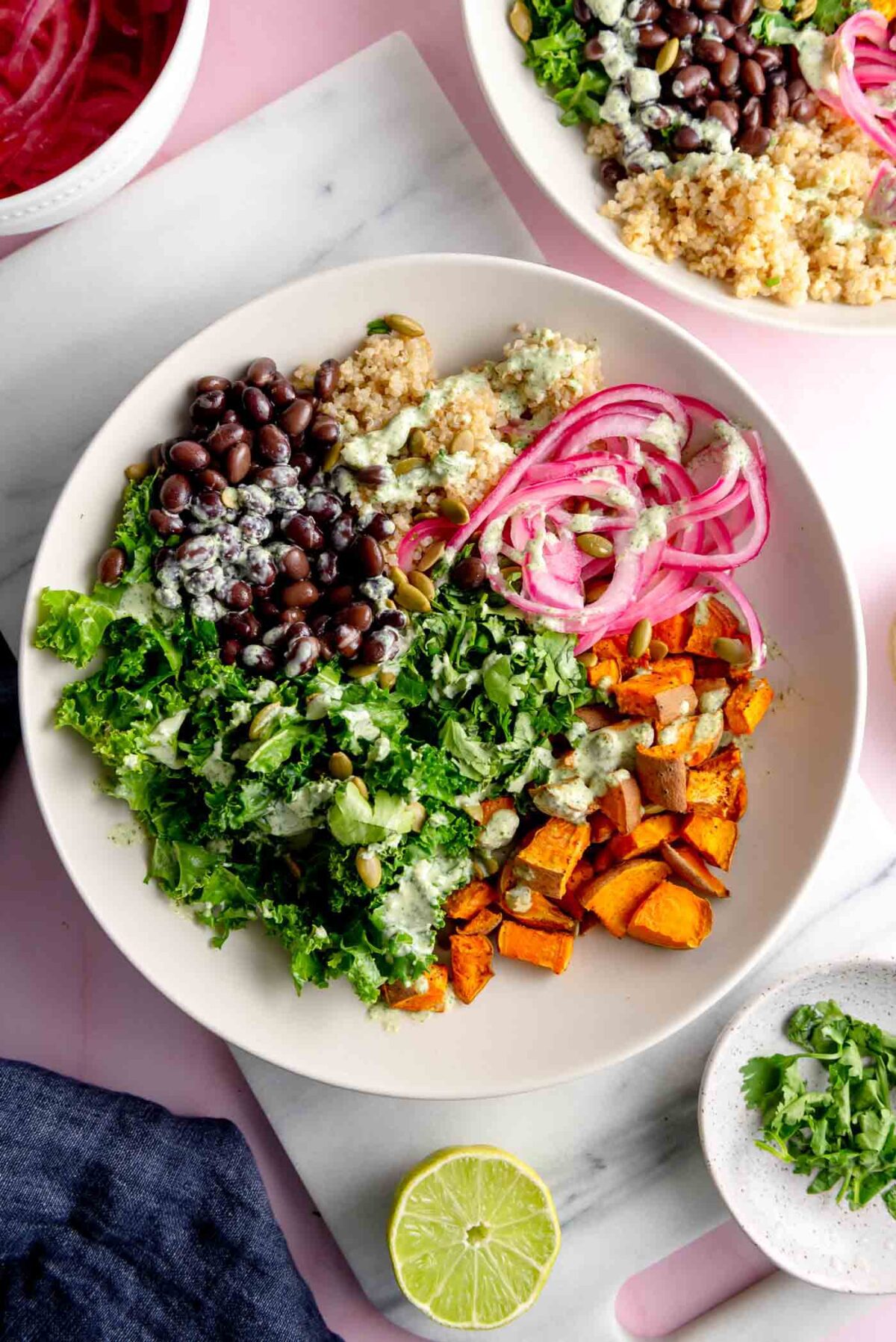 Bowl on a marble cutting board with roasted sweet potato, kale, black beans, quinoa, thinly sliced red onion and a creamy green sauce on top and a sliced lime beside the bowl.