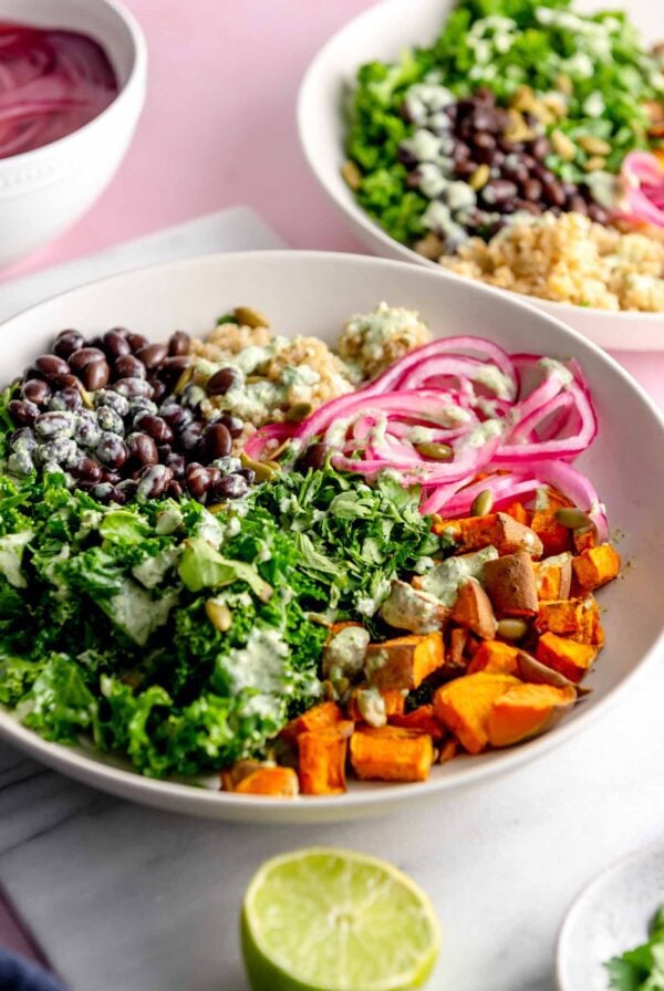 Bowl on a marble cutting board with roasted sweet potato, kale, black beans, quinoa, thinly sliced red onion and a creamy green sauce on top and a sliced lime beside the bowl.