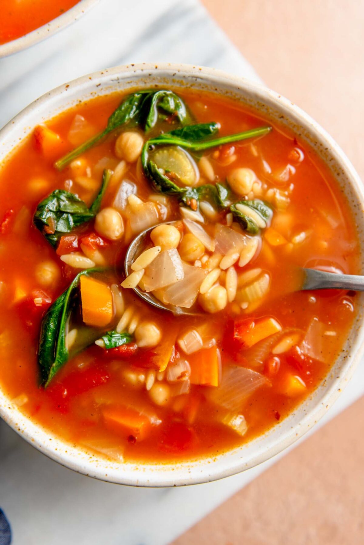 Bowl of tomato orzo soup with spinach and chickpeas with a spoon in it on a cutting board.