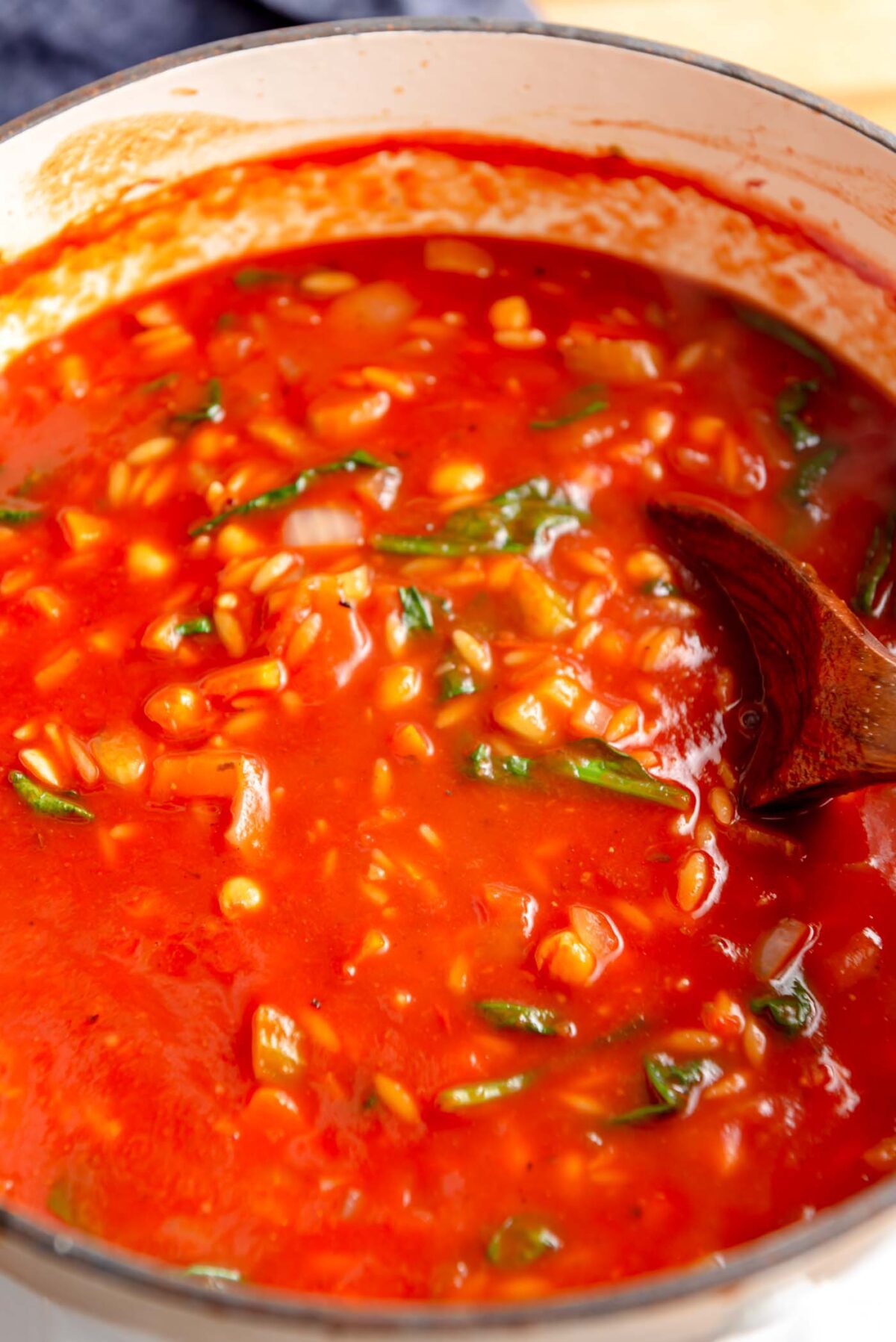 Tomato orzo and spinach soup cooking in a soup pot with a wooden spoon.