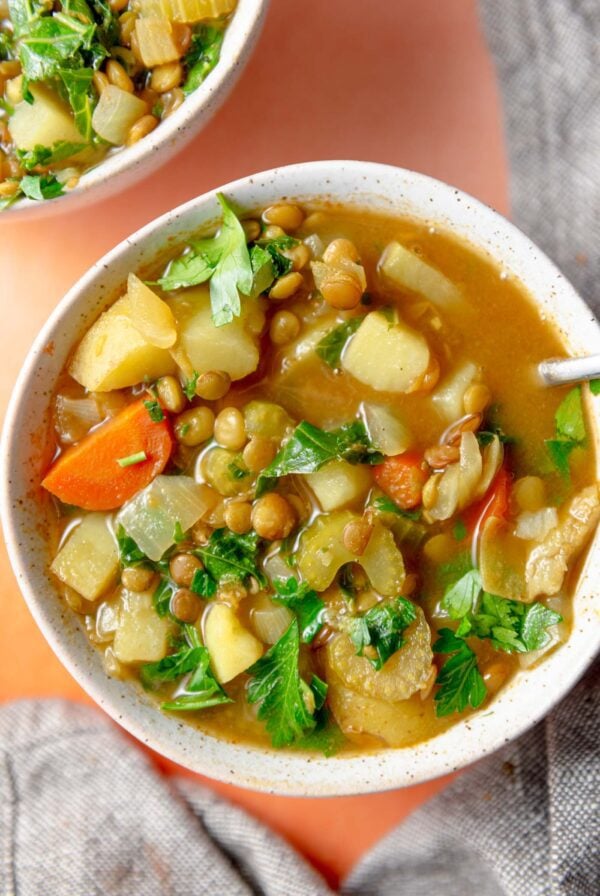 Bowl of a hearty soup with potatoes, carrots, lentils and kale topped with kale with a spoon resting in it.