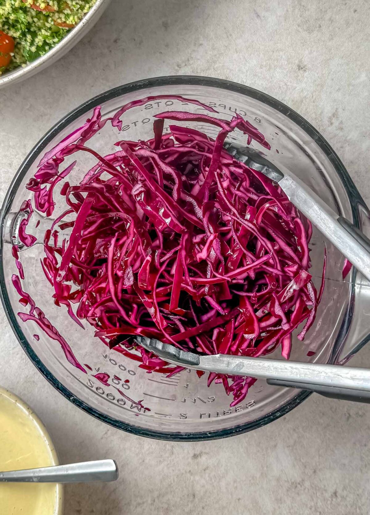 Shredded cabbage in a glass bowl with a pair of tongs in it. 