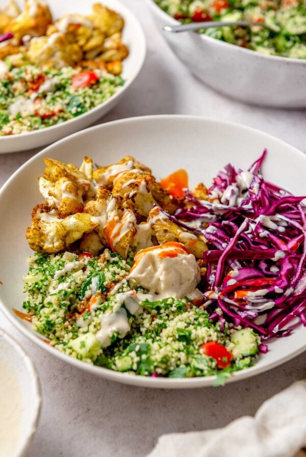 Cauliflower shawarma bowl with roasted spiced cauliflower, shredded cabbage, couscous tabouleh, hummus and tahini sauce and hot sauce drizzled over top.