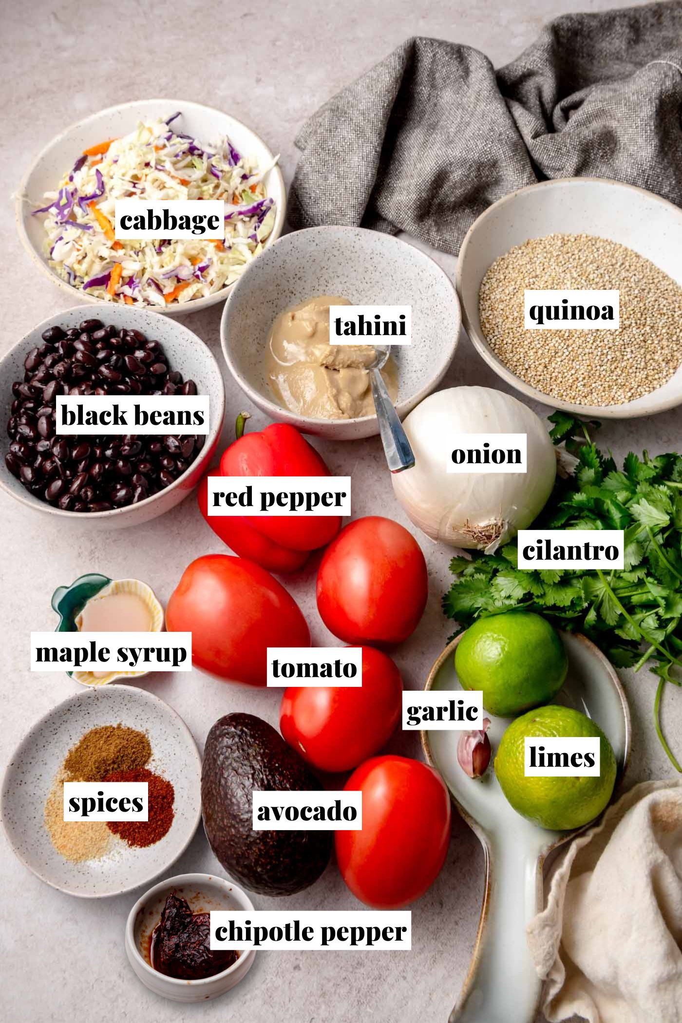 Flat lay of all ingredients for a chipotle black bean quinoa bowl, including bowls of shredded cabbage, cooked black beans, tahini, quinoa, maple syrup, spices, and chipotle pepper, arranged alongside whole tomatoes, red pepper, avocado, onion, garlic, limes, and fresh cilantro. Each ingredient is labeled with text overlays.