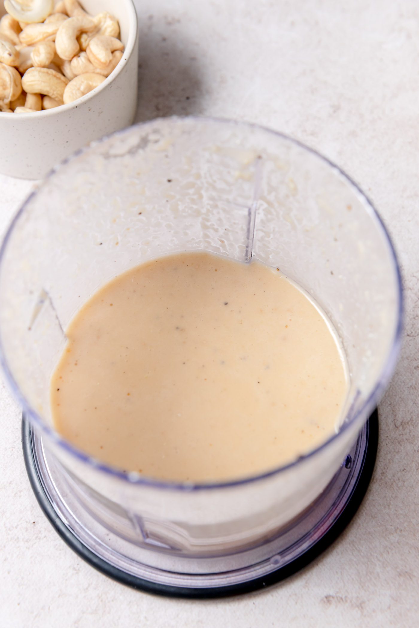 A blender cup filled with creamy miso ginger sesame dressing, pale beige in color with tiny specks of seasoning. A small bowl of raw cashews sits nearby on the countertop.
