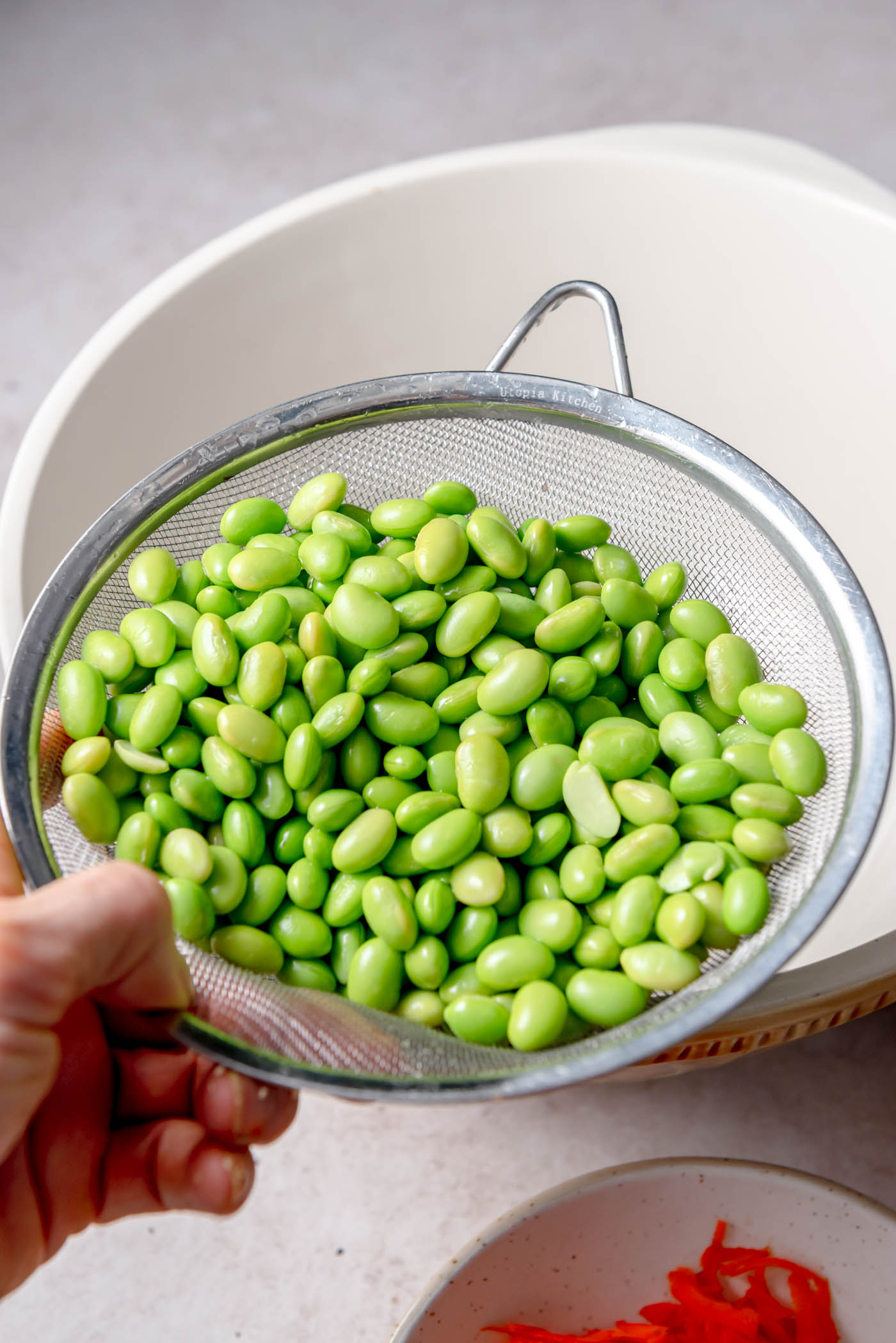 A metal strainer filled with freshly rinsed bright green edamame beans held over a large white mixing bowl, ready to be added to the salad.