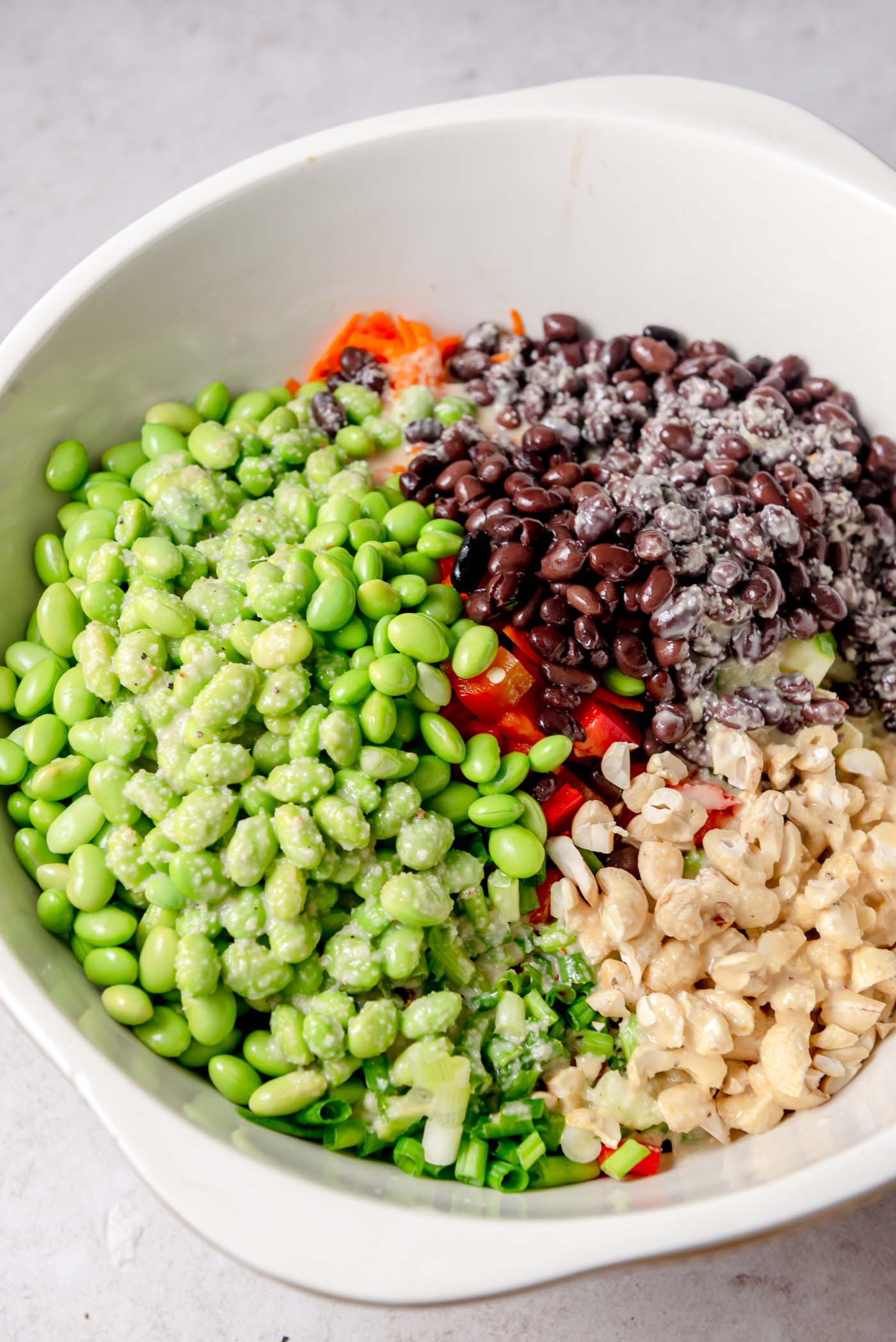 A large white mixing bowl containing ingredients for the edamame bean salad: edamame, black beans, chopped red pepper, cashews, and green onions, arranged in sections before being mixed together.