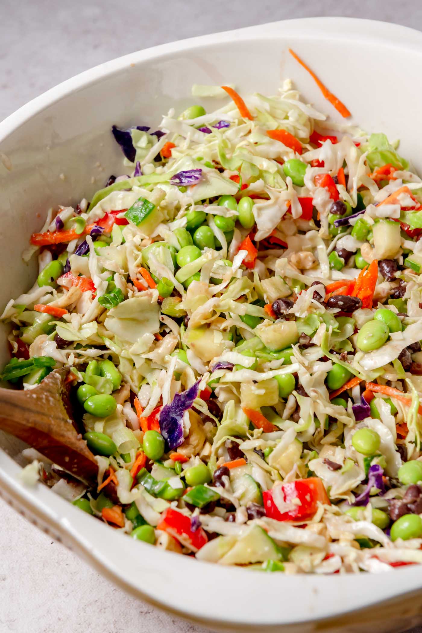 Edamame bean salad ingredients tossed together in the mixing bowl, cabbage, edamame, black beans, red pepper, carrot, cucumber, and green onions coated lightly with dressing, creating a colorful mix.