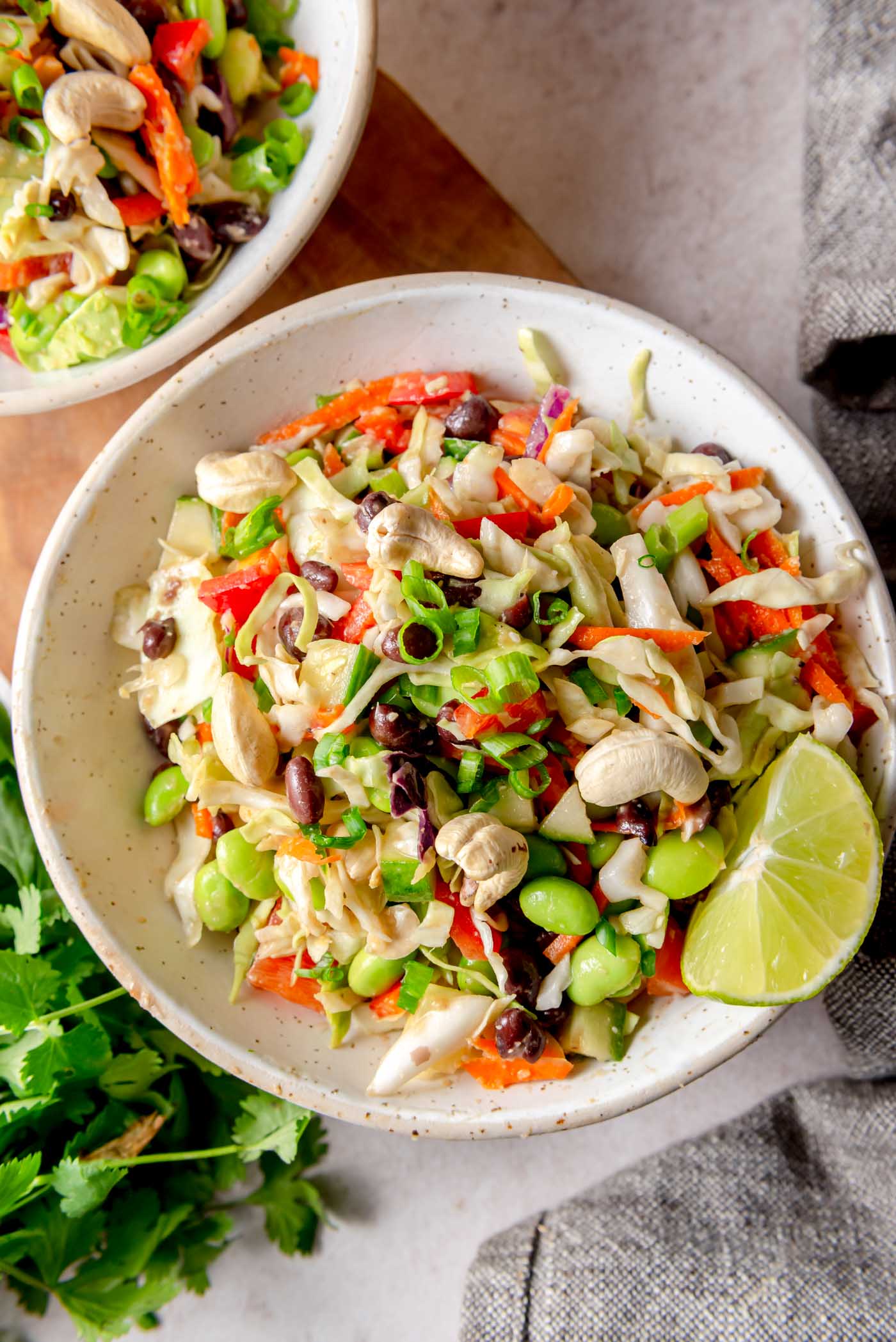 Top-down view of two bowls of finished edamame bean salad topped with cashews and chopped green onions, served with a wedge of lime and fresh cilantro on the side.