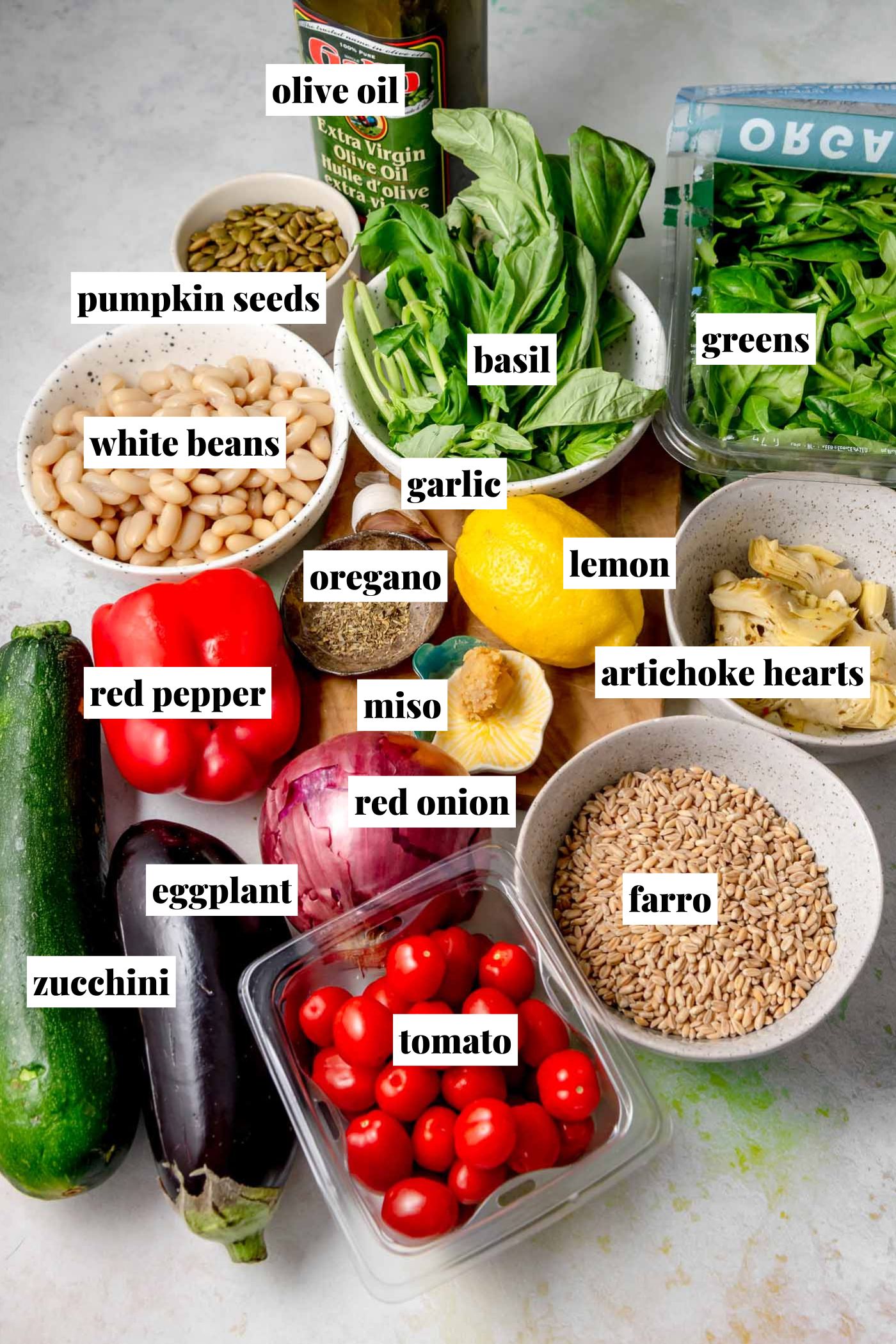 Labeled overhead photo of all ingredients for a warm pesto farro salad, including fresh basil, greens, olive oil, lemon, garlic, oregano, miso, white beans, pumpkin seeds, artichoke hearts, farro, cherry tomatoes, red pepper, red onion, zucchini, and eggplant arranged on a countertop.