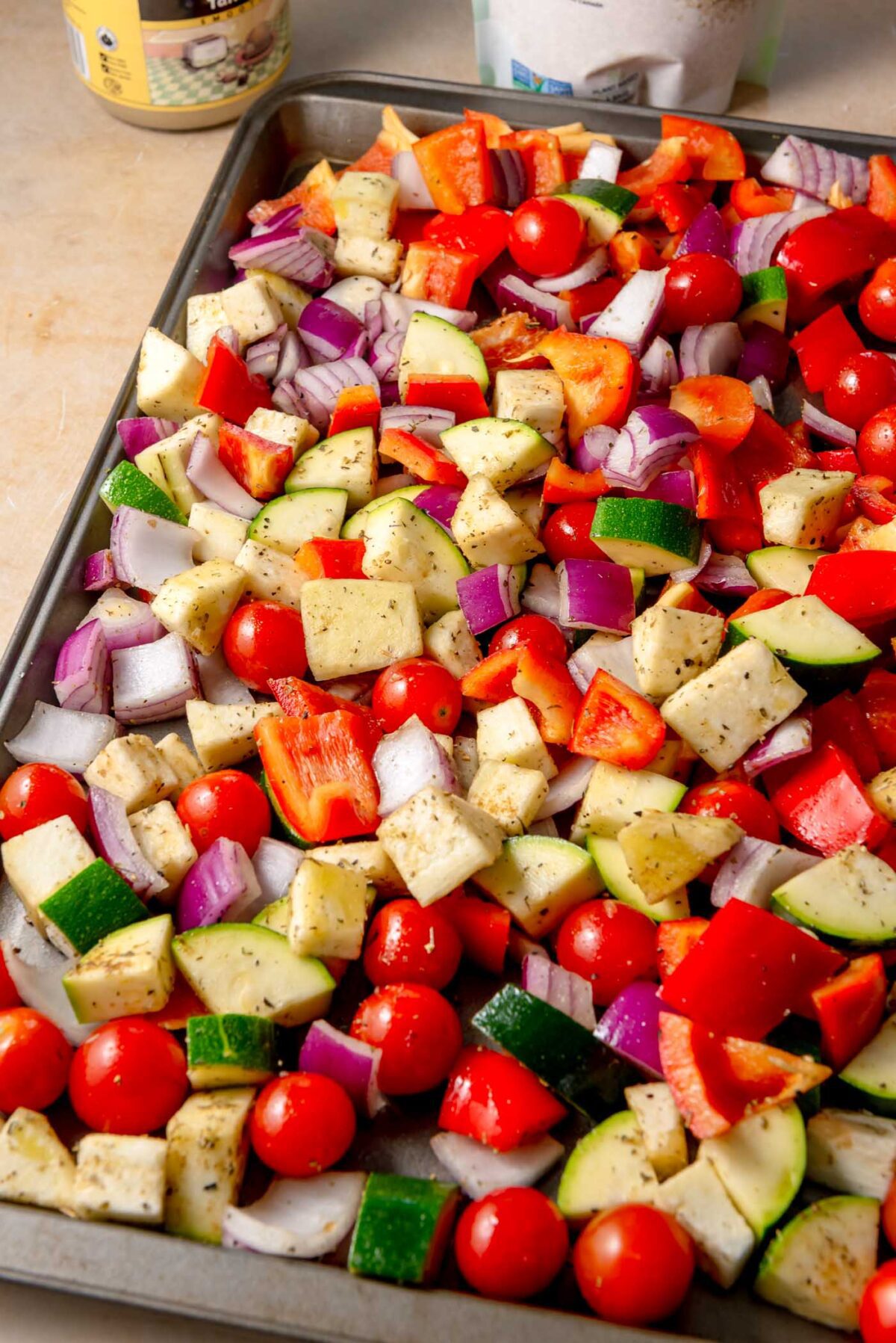 Colorful chopped zucchini, red peppers, eggplant, onion, and cherry tomatoes on a sheet pan seasoned with herbs.