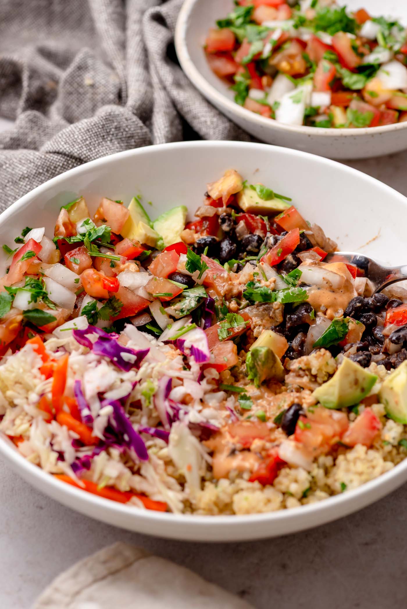 Close-up of a mixed burrito bowl showing pico de gallo, avocado, black beans, slaw, quinoa, and chipotle sauce.