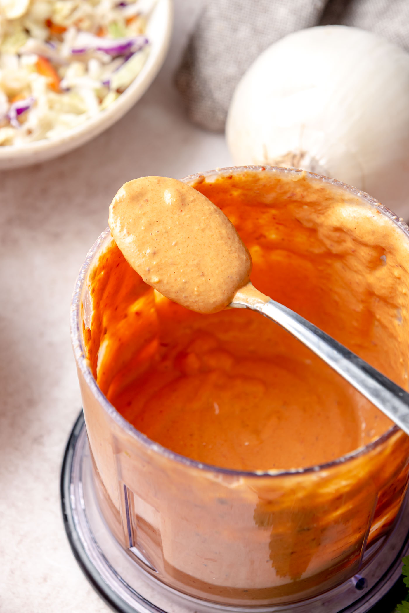 A close-up of a spoon lifting thick, creamy chipotle-style sauce from a food processor bowl.